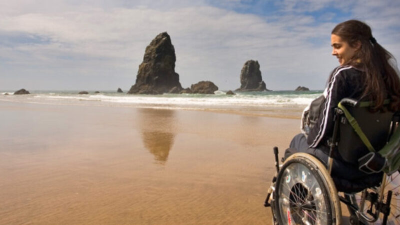 Young woman in a wheelchair smiling while looking out at the ocean on a sandy beach, with large sea stacks in the distance under a partly cloudy sky.